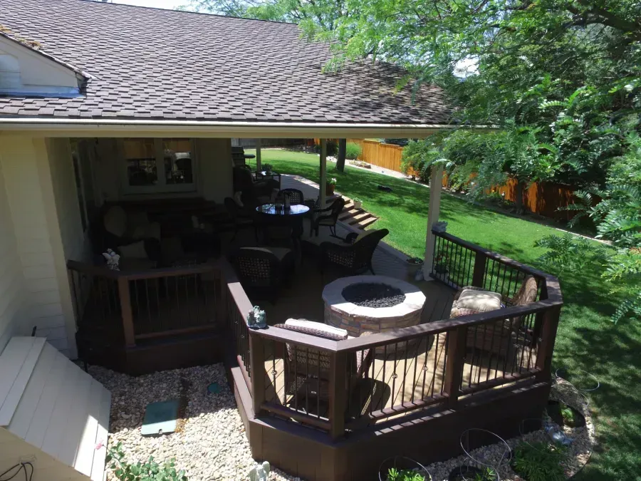 Covered wooden deck with patio furniture and a round table beside a house, overlooking a green yard