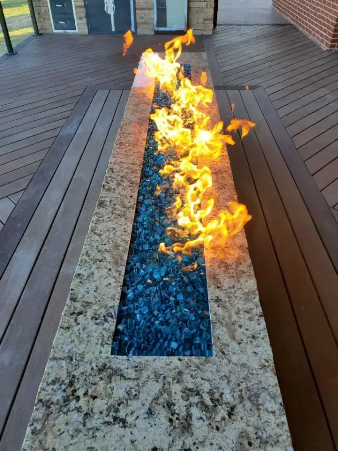 Stone fire pit with bright flames burning along a narrow blue glass channel on a wooden deck