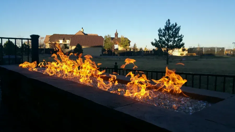 Outdoor fire pit with bright flames along a dark raised edge at dusk, near a grassy field and houses.