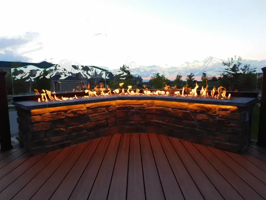 Curved stone fire pit with flames on a wooden deck at dusk, mountain view in the background