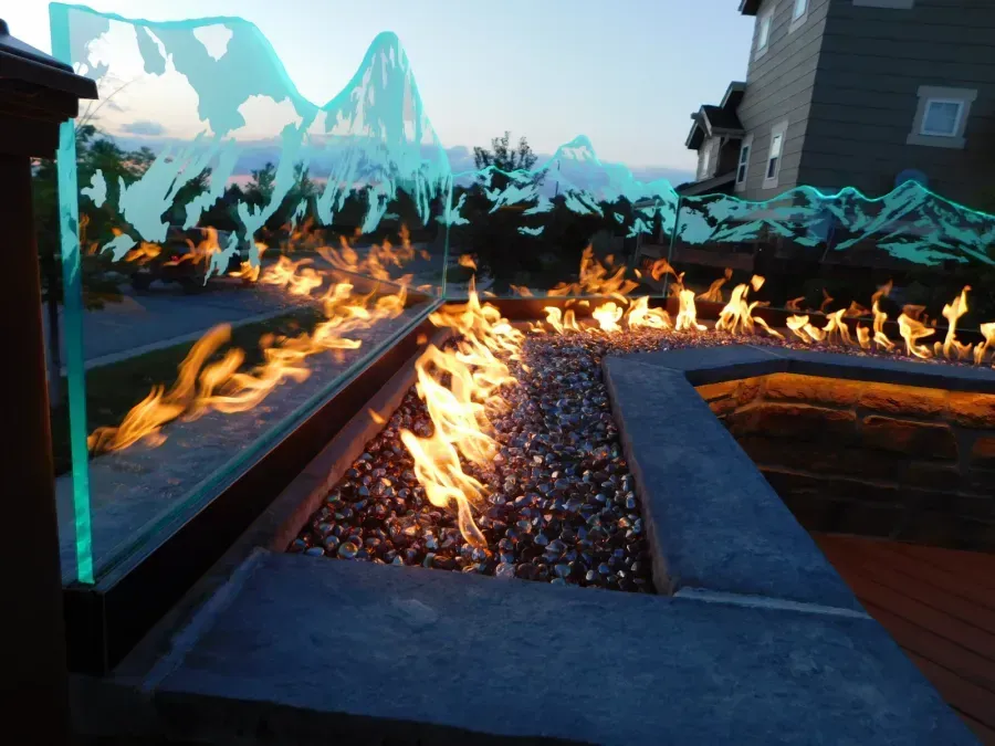 Modern backyard fire pit with flames along a gravel-lined water feature beside a deck at dusk