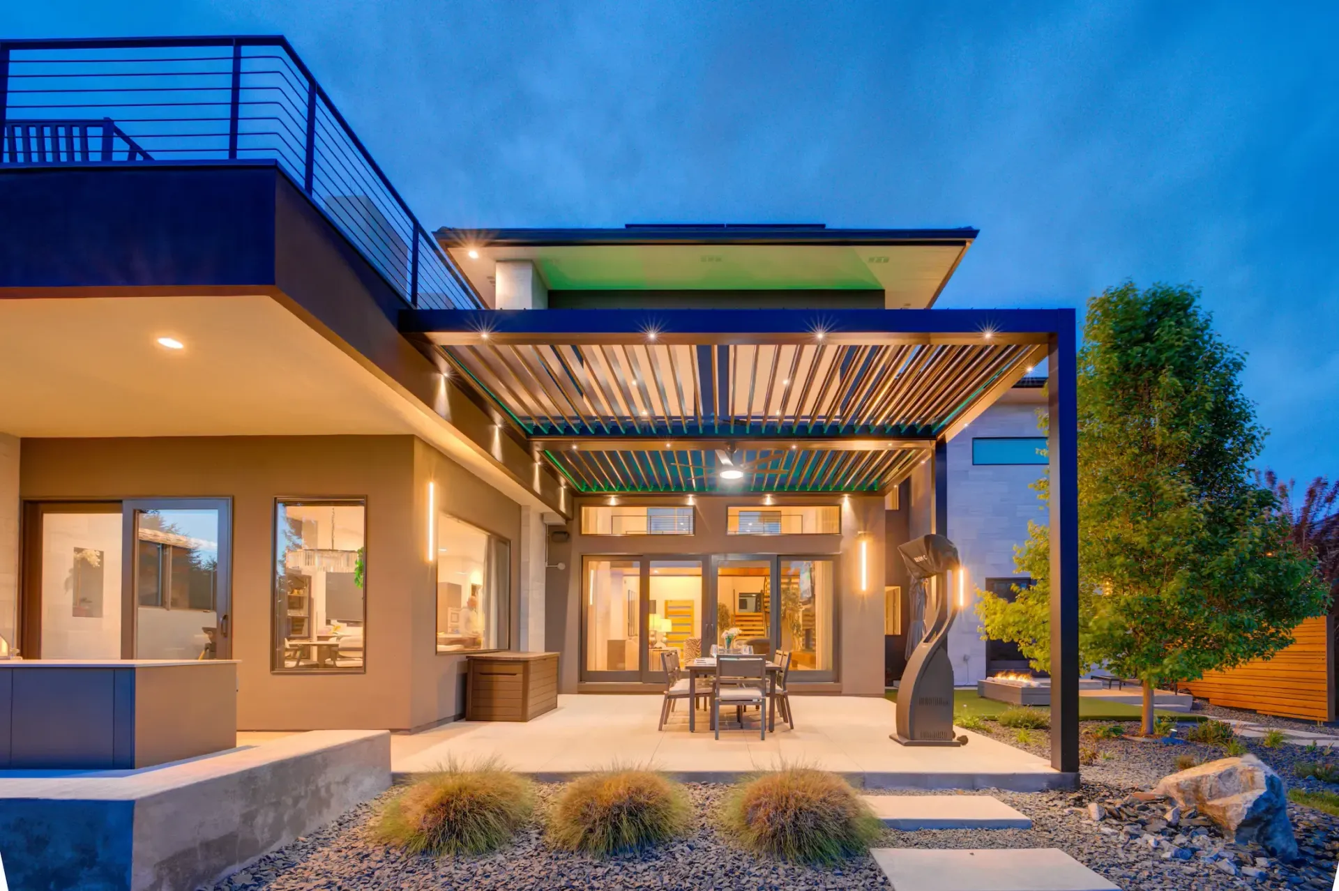 Modern house patio at dusk with pergola, warm interior lights, and desert landscaping