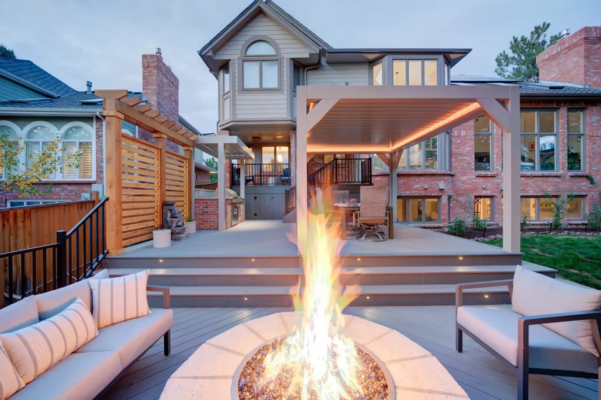Modern house patio with fire pit, cushioned seating, wooden pergola, and warm evening lighting