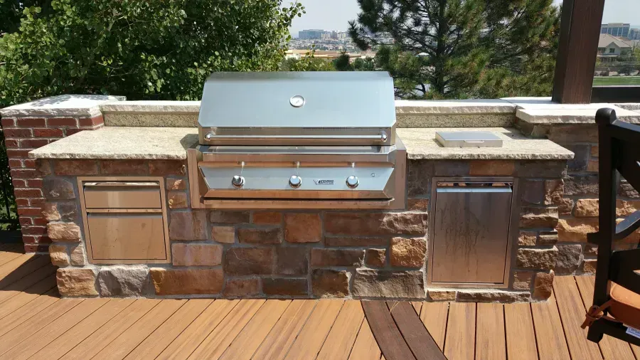 Outdoor stone barbecue grill with stainless steel lid and side drawers on a wooden deck