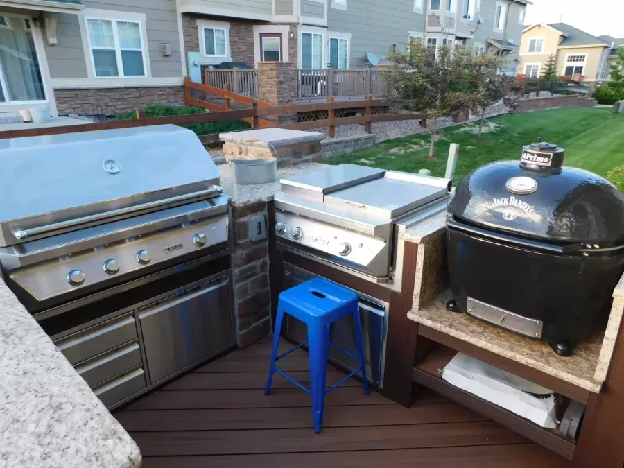 Outdoor patio with stainless grill, black smoker, and blue stool beside a stone counter near houses.