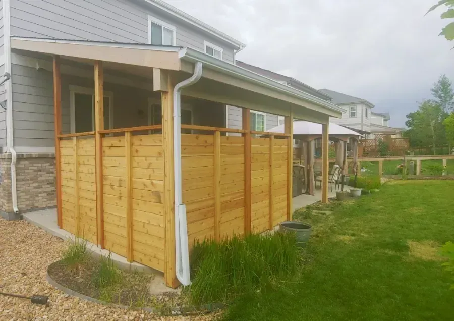 Covered wooden patio enclosure beside a gray house with a sloped roof and green lawn