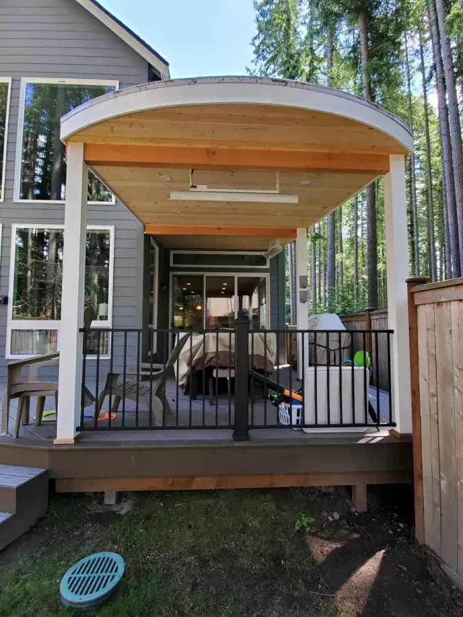 Covered deck with black railing and outdoor chairs beside a modern house in a wooded yard