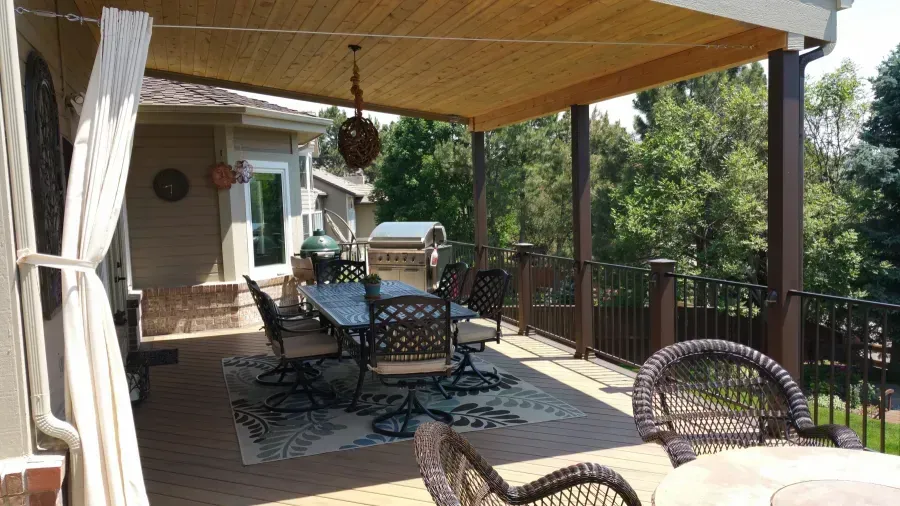 Covered patio with wicker table and chairs, wooden ceiling, and a view of trees and a house entry.