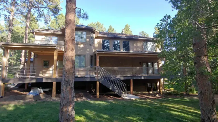 Wooden house with large balconies and stairs, surrounded by tall pine trees and a grassy yard.
