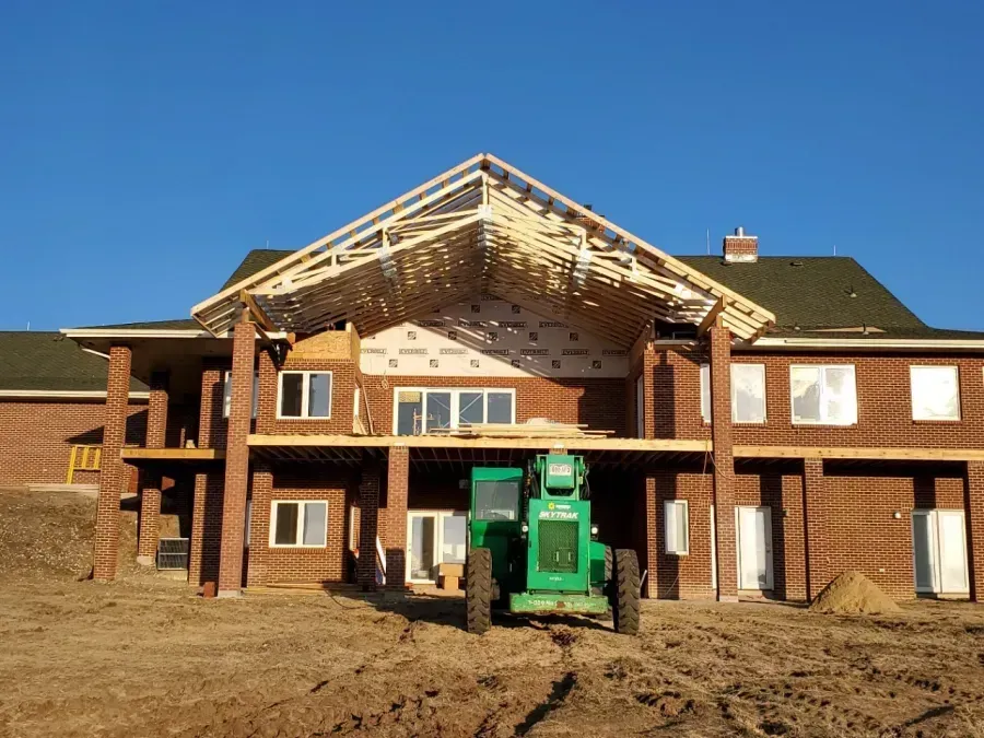 Brick house under construction with exposed roof trusses and a green front loader in the dirt yard