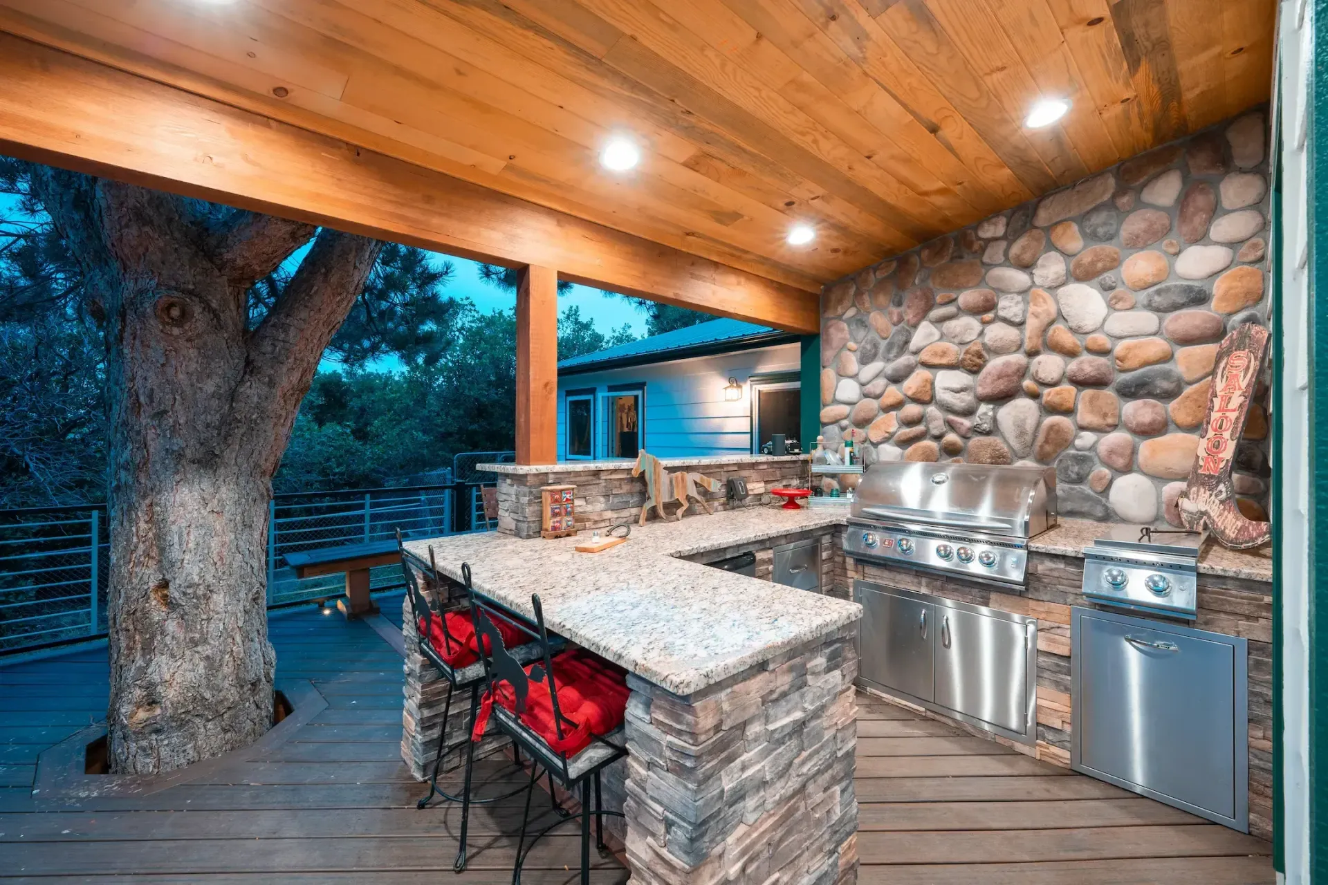 Covered outdoor kitchen with granite island, stone wall, built-in grill, and wood ceiling at dusk