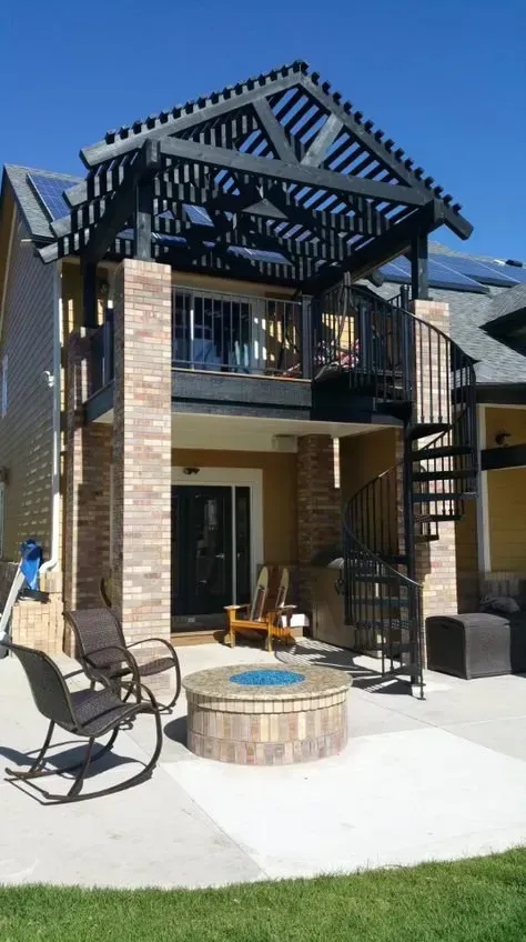 Backyard patio with pergola, balcony, seating, and a round fire pit under a two-story house.