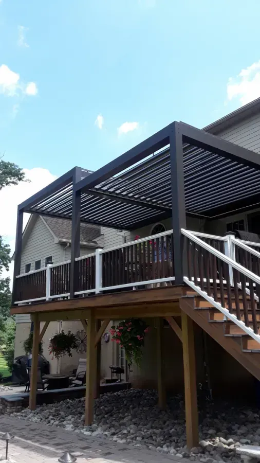 Elevated wooden deck with dark pergola roof and stairs beside a house under a bright sky