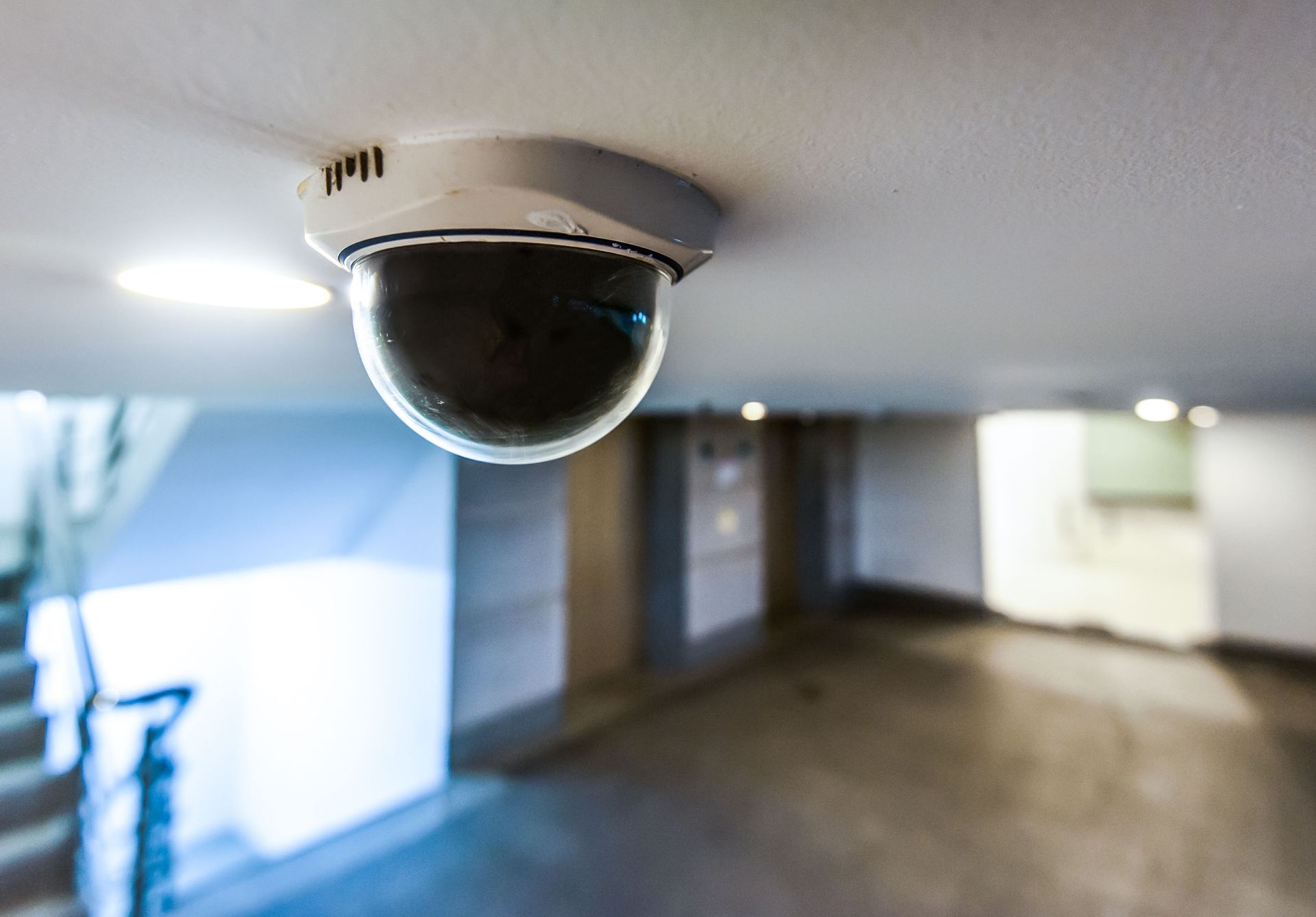 Security camera on a white ceiling, overlooking a brightly lit hallway.