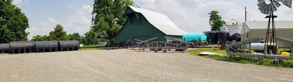 A large green barn is sitting in the middle of a gravel driveway next to a windmill.