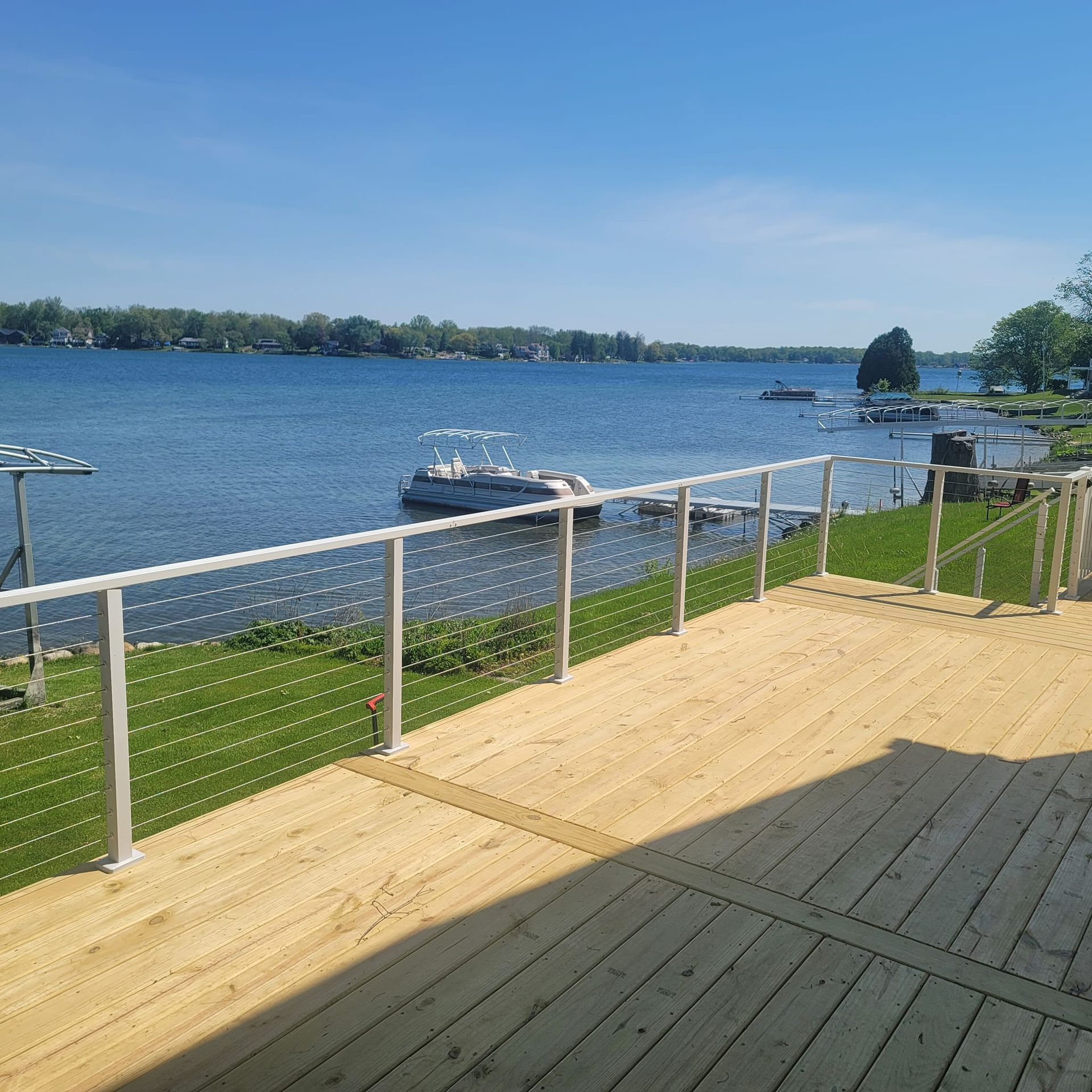 Wooden deck overlooking a lake with boats, blue sky, and green grass.