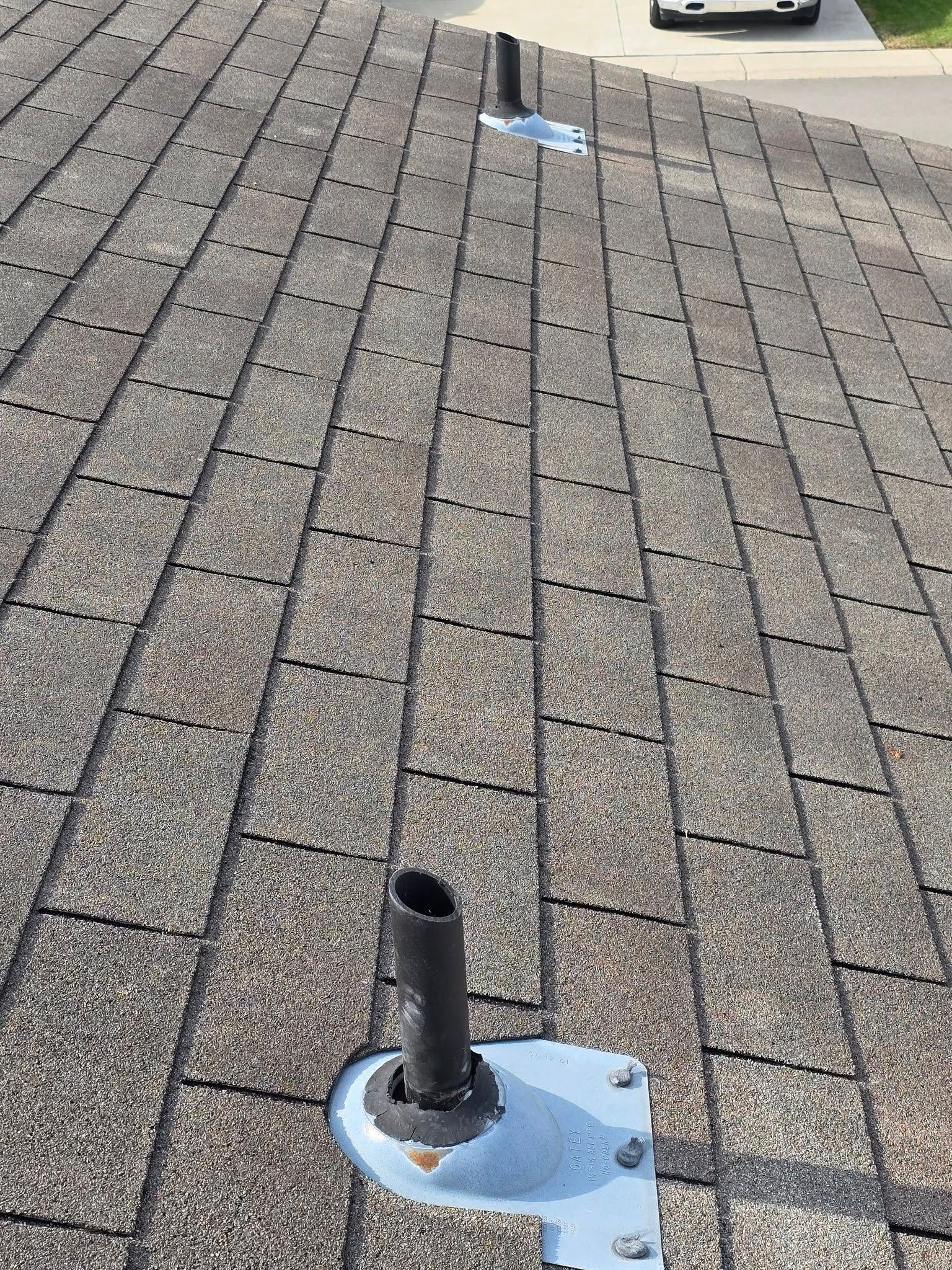 Black pipes on a weathered asphalt shingle roof with metal flashing. Sunny day.