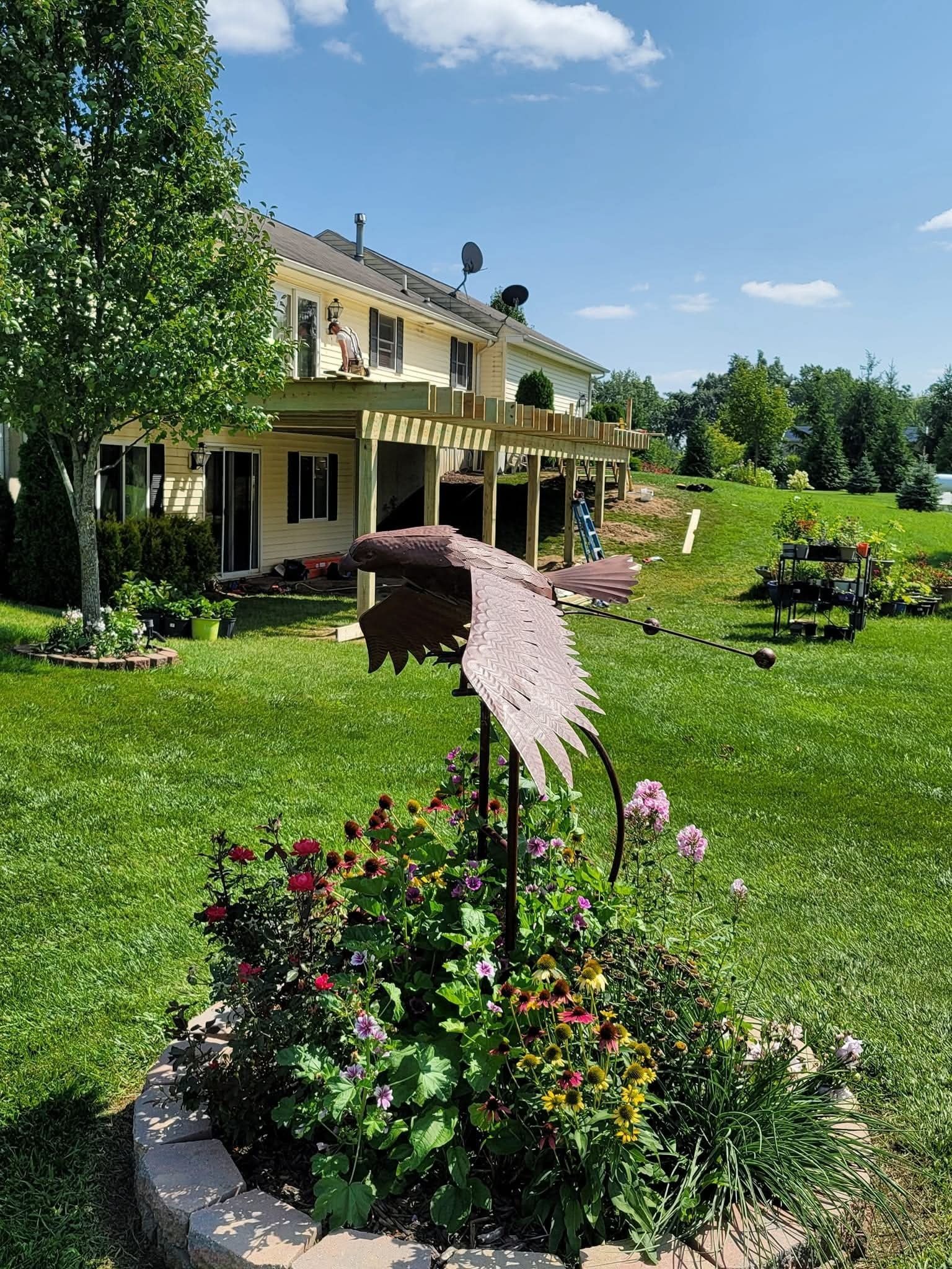Backyard with house, wooden deck, and metal art sculpture surrounded by flowers. Sunny day, green lawn.