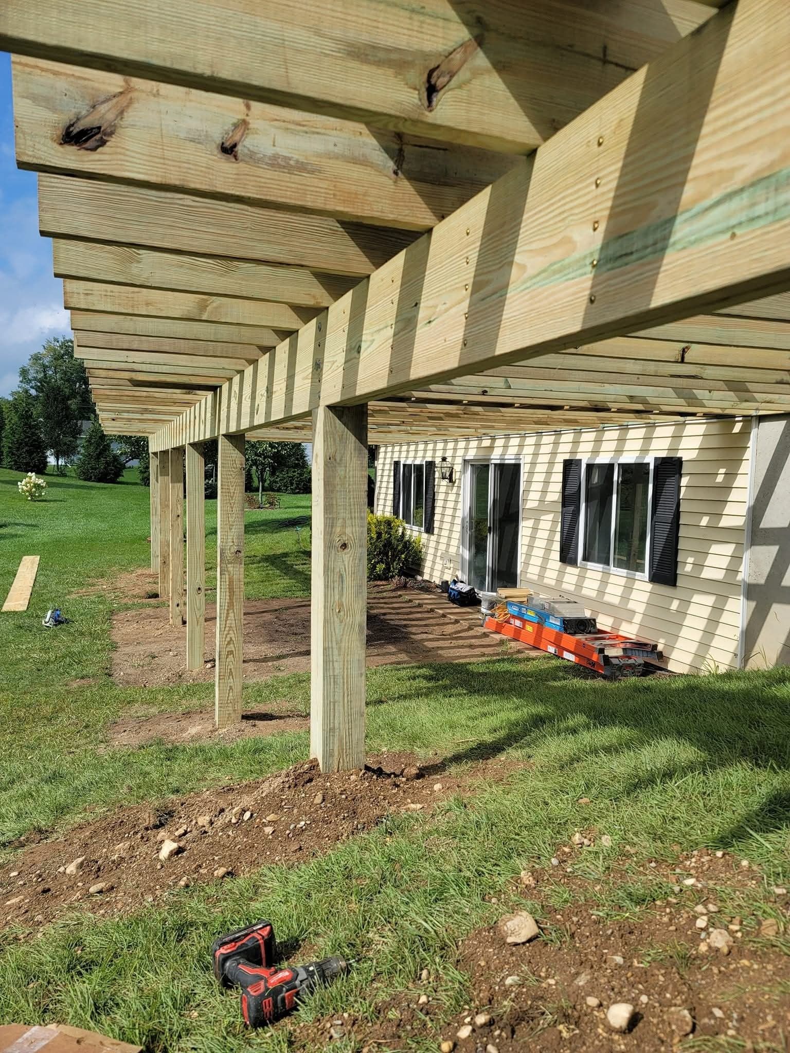 Deck construction with wooden beams, posts, and boards. Green grass and a house in the background.