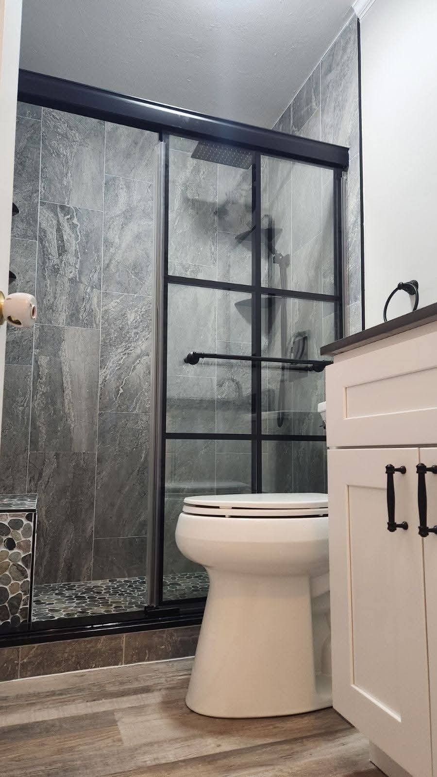 Modern bathroom with a black-framed shower, white toilet, and vanity. Gray tile and wood-look flooring.