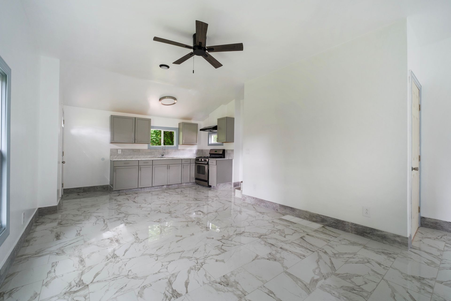 An empty room with a ceiling fan and a kitchen in the background.