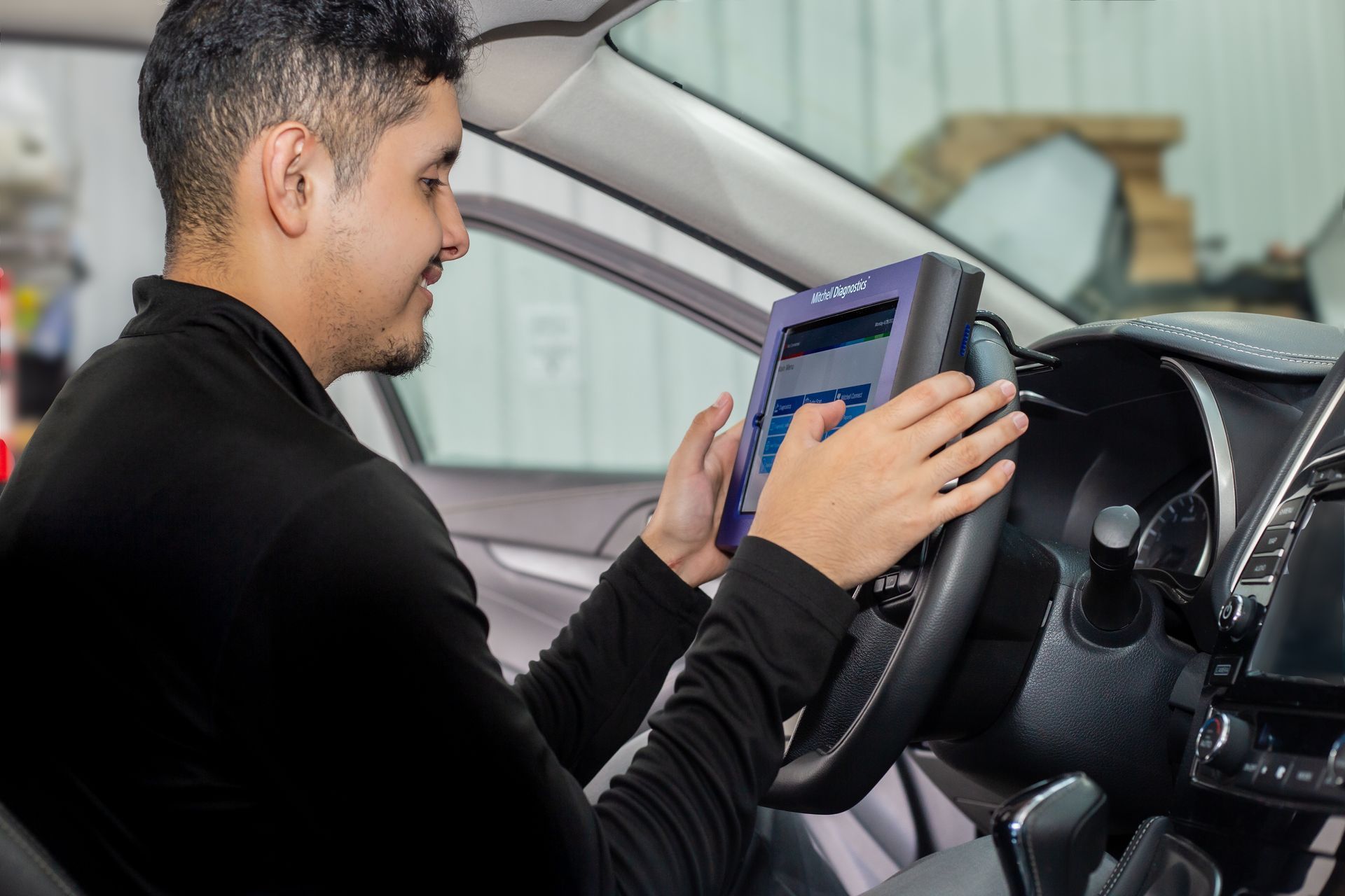 A technician in a black long-sleeve shirt uses a diagnostic tablet while seated in a car, checking vehicle systems.