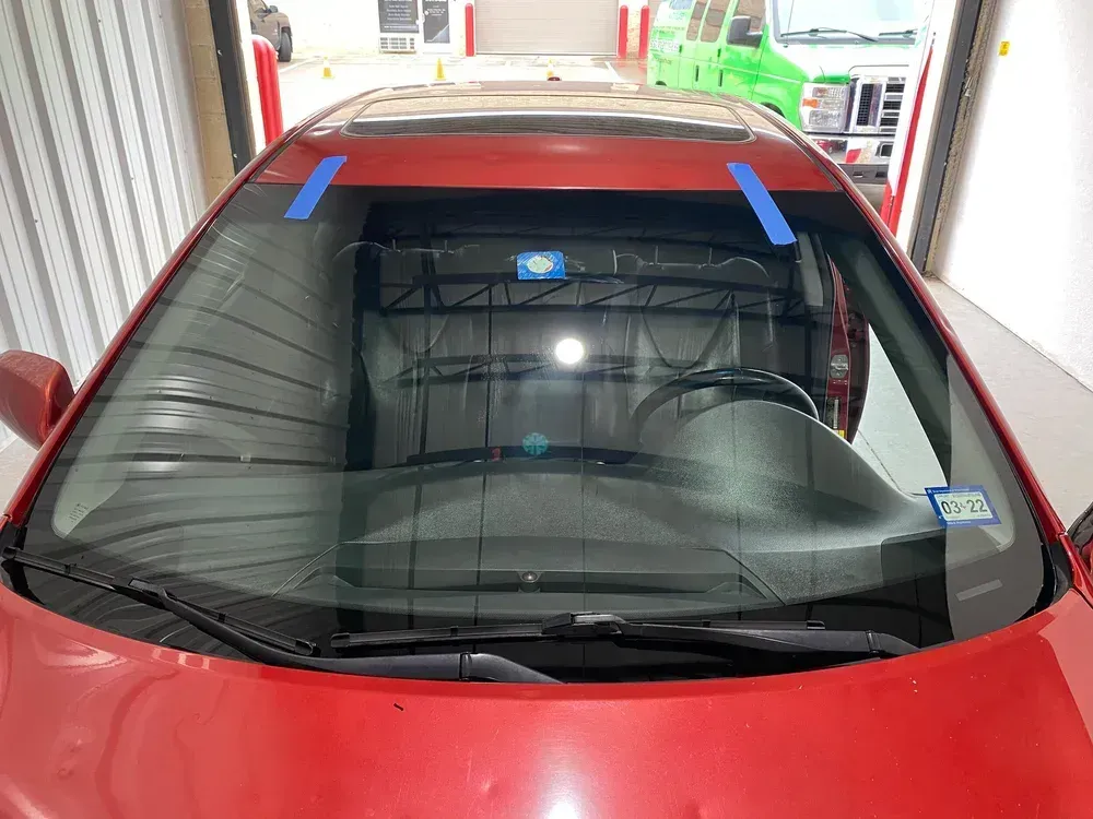 Red car with windshield taped for repair in a garage.