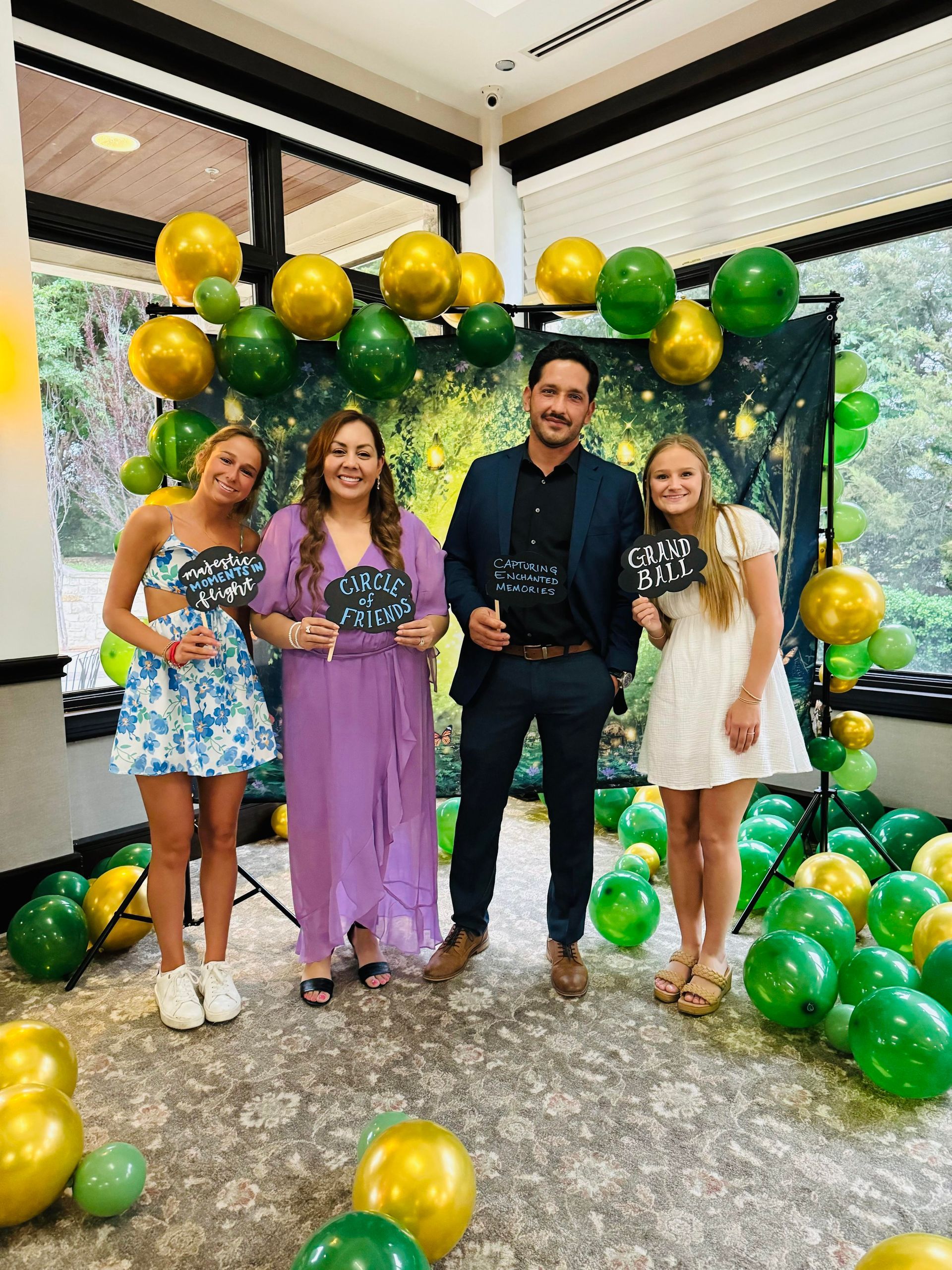Four people pose with props, surrounded by green and gold balloons, smiling.