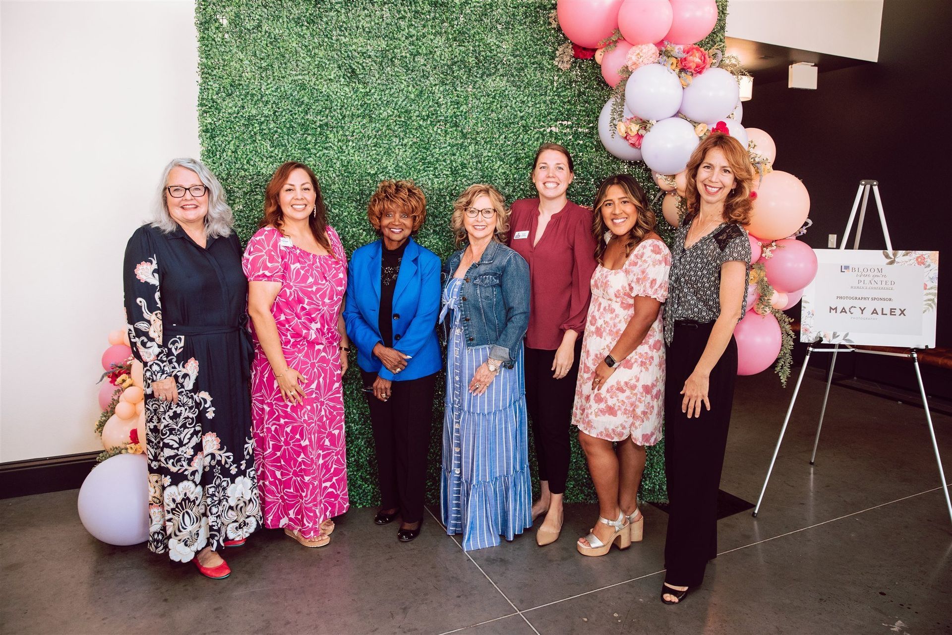Group of eight women posing in front of a greenery wall and balloons, smiling.