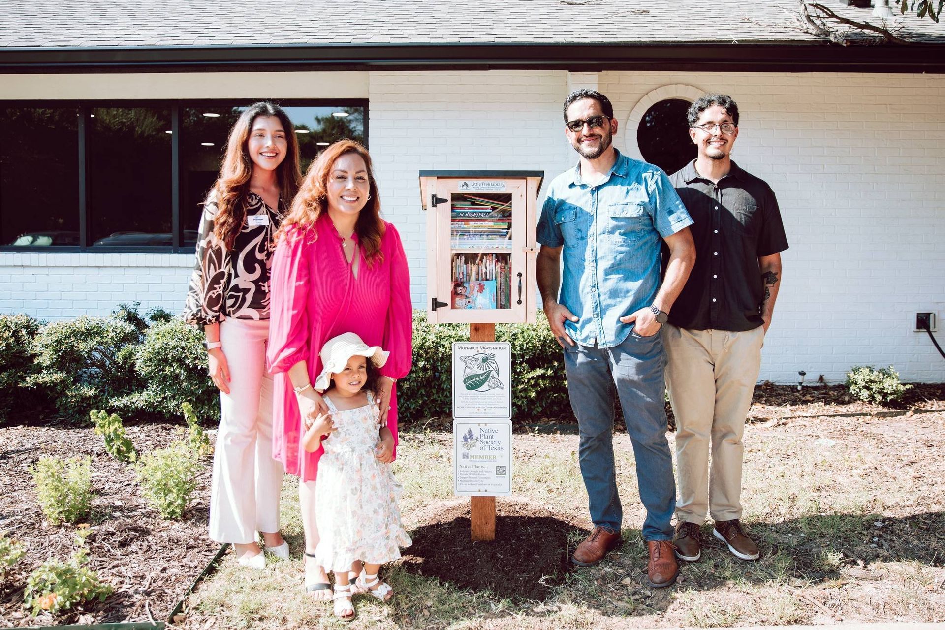 Family poses in front of a Little Free Library. The woman in pink holds a child, others smile.