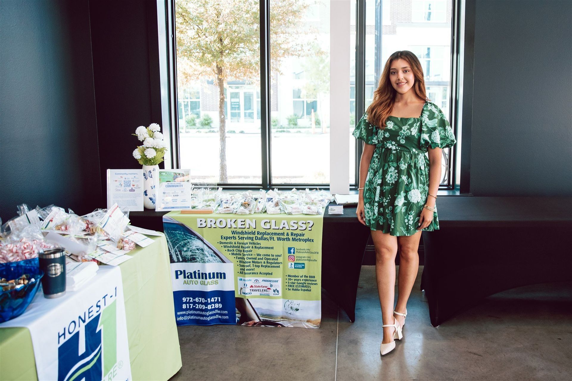 Woman in green dress stands by a table with products. Window and outdoor view visible.