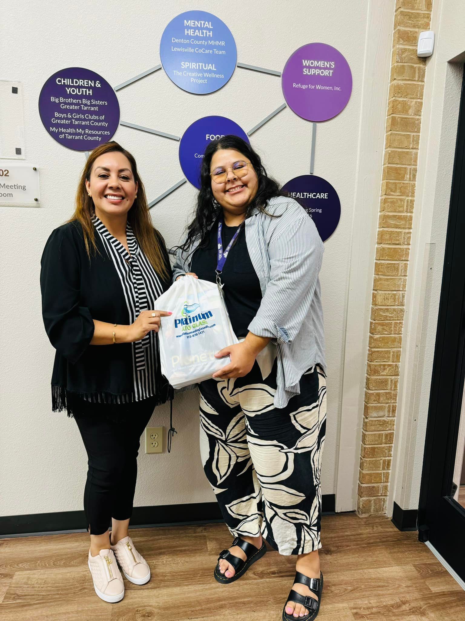 Two women holding a gift bag, smiling, in front of a wall with circles and text.