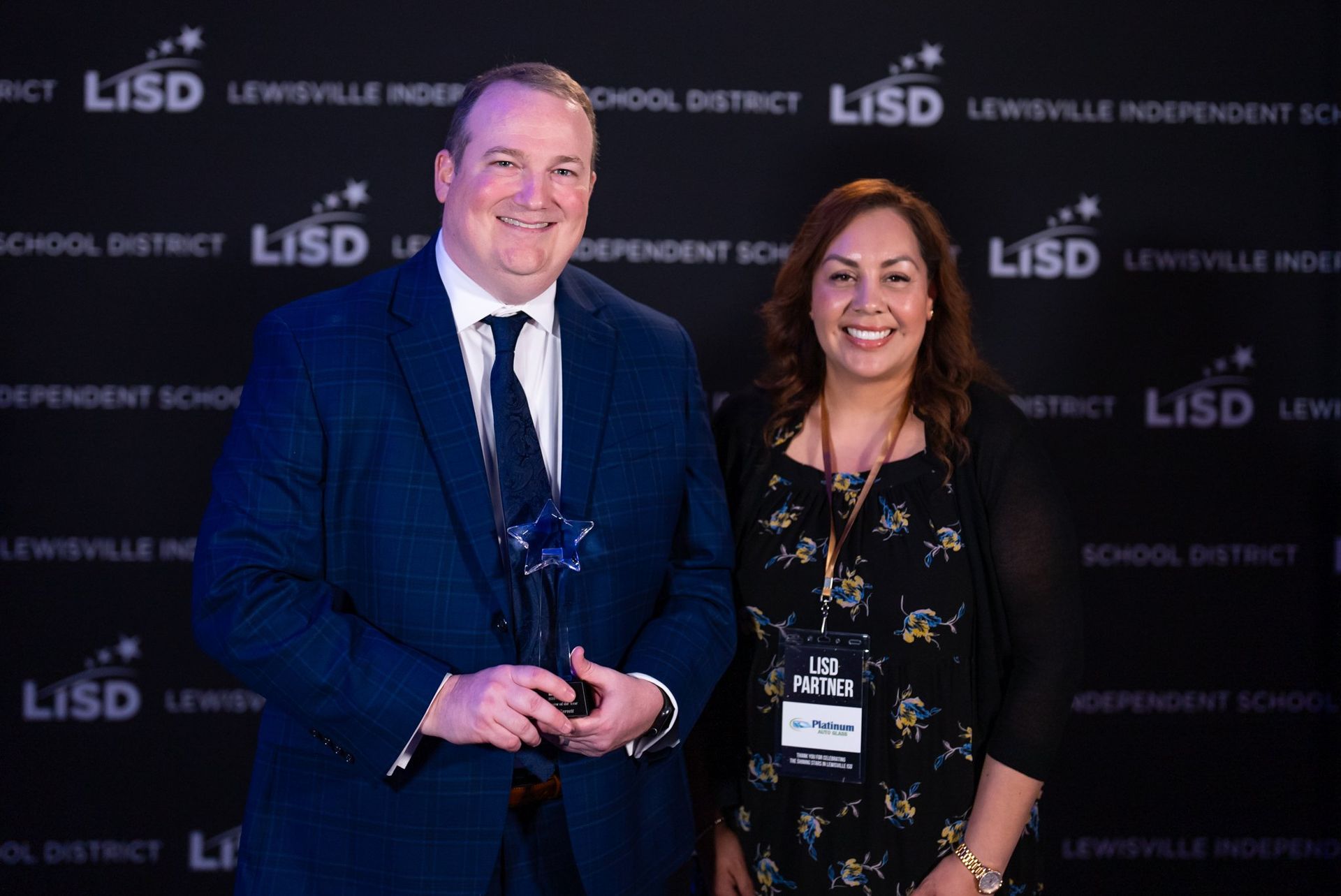 Man in suit holds award next to woman with name tag; Lewisville ISD backdrop.