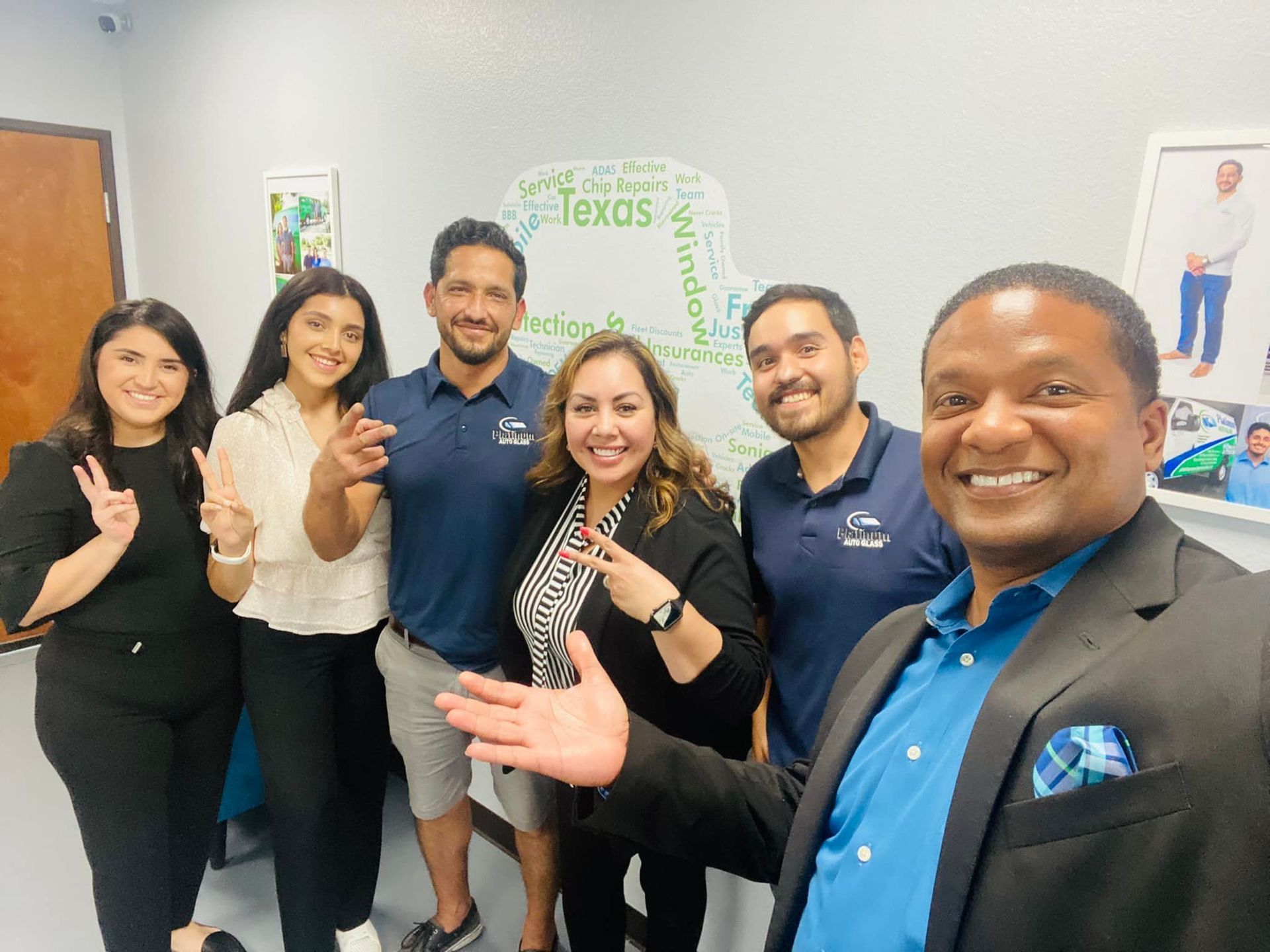 Group of smiling people posing in front of a Texas-themed wall. Some are giving peace signs.