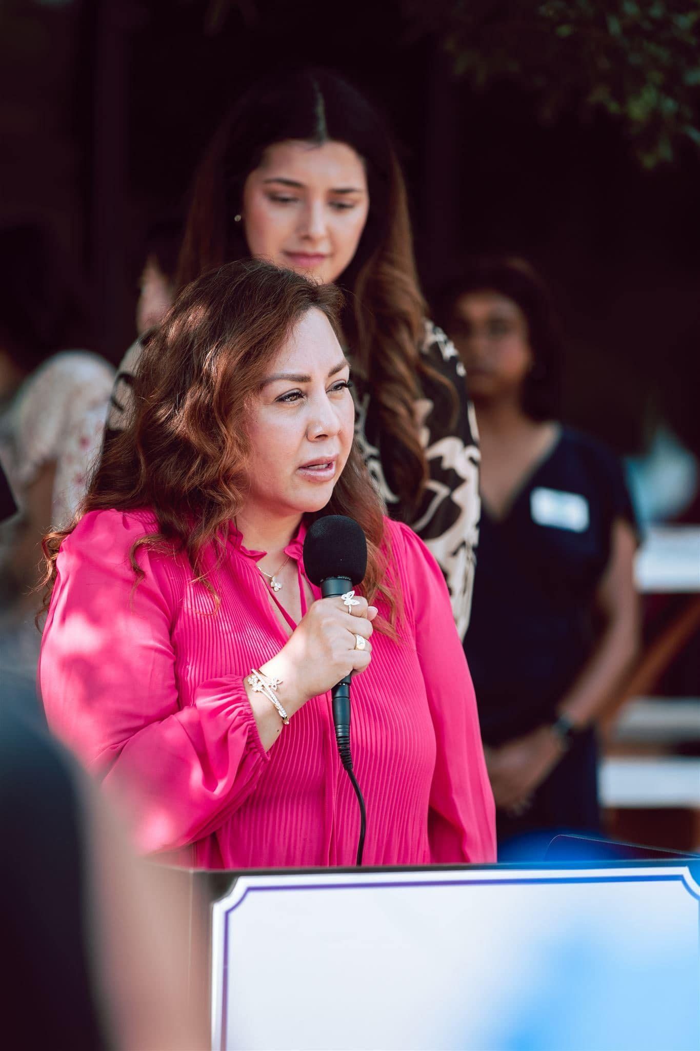 Woman in pink shirt speaks into a microphone. Two women stand behind her outdoors.