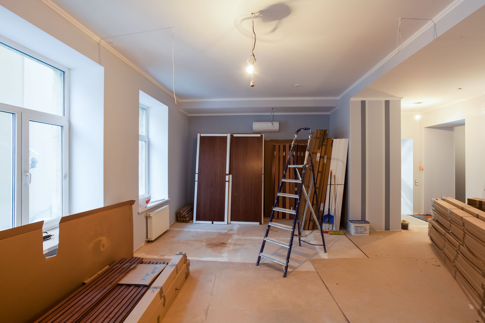 Renovation in progress in an empty room with ladders, stacked materials, and exposed fixtures.