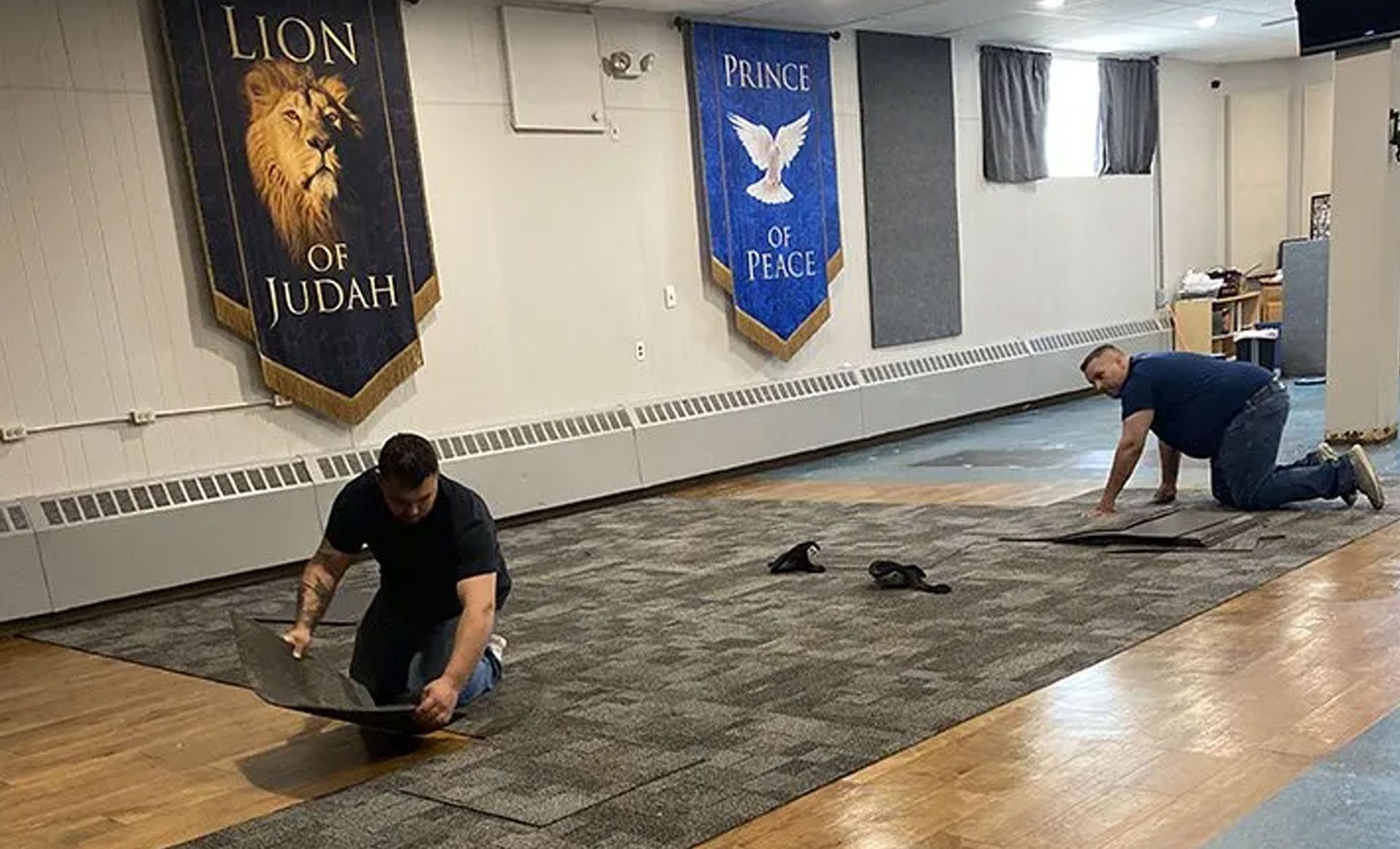 Two people kneeling on mats in a studio, stretching or exercising on a wooden floor with banners on the wall