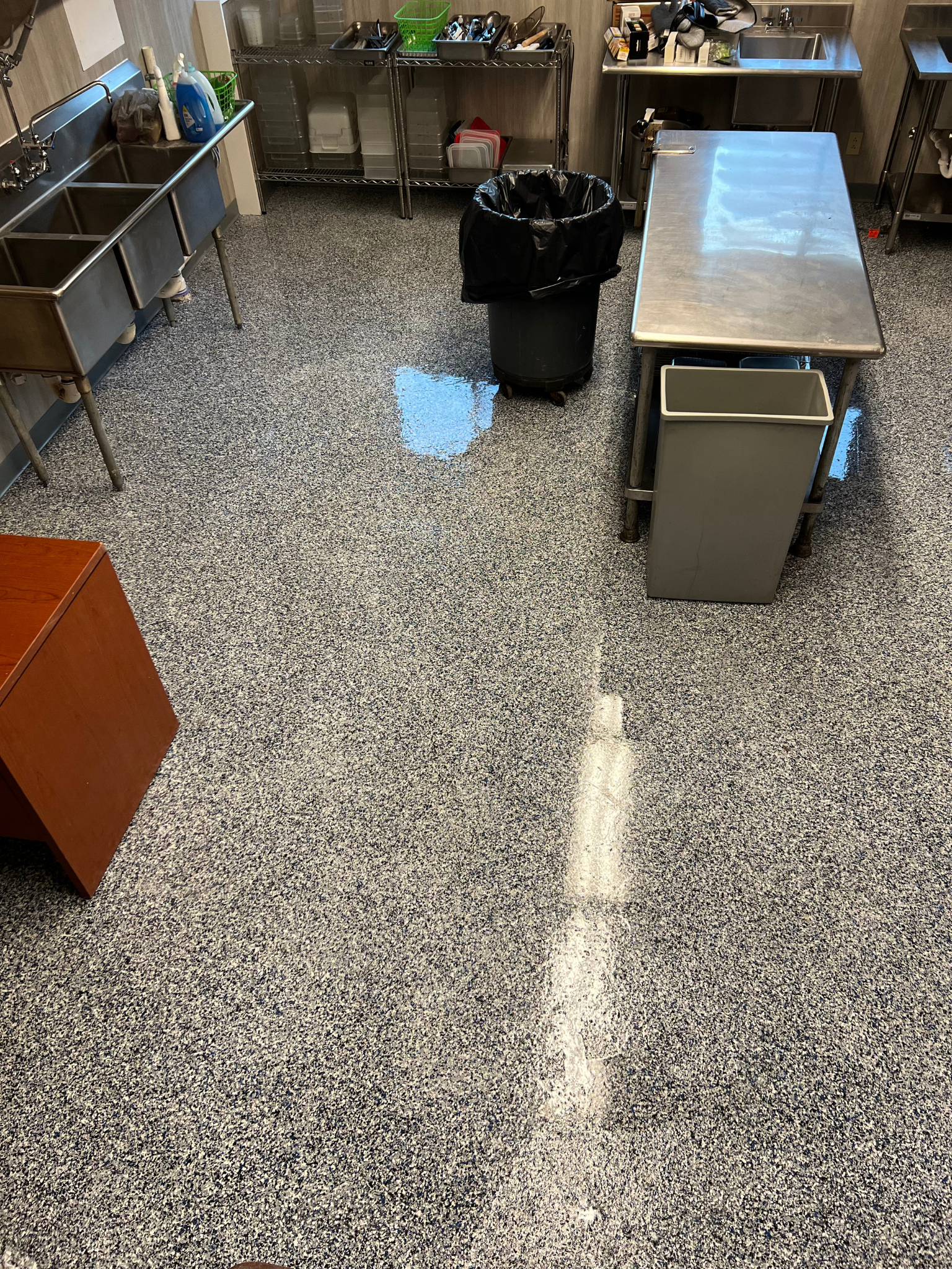 Small commercial kitchen with metal prep tables, sink, trash can, and speckled floor.