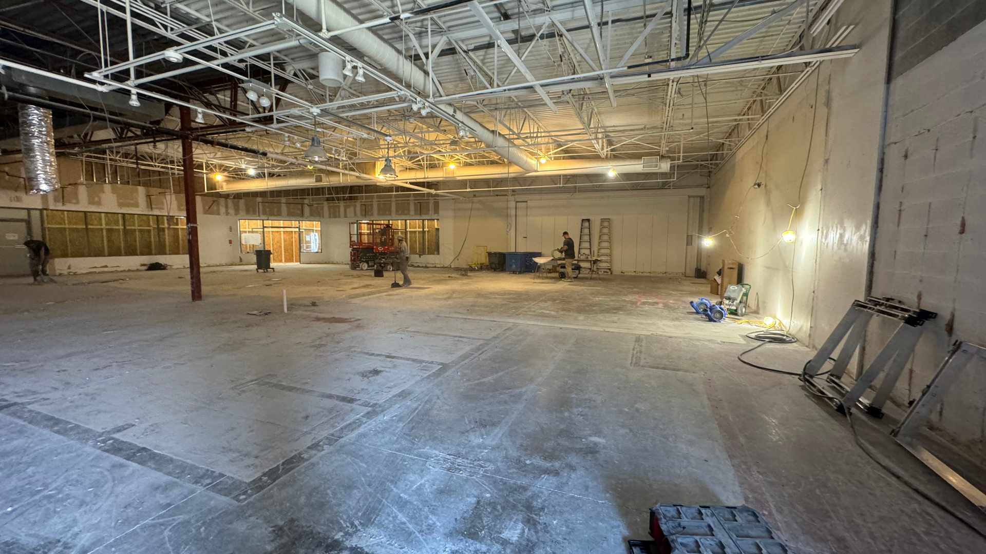 Empty warehouse interior under renovation with exposed ceiling, concrete floor, and construction equipment along the walls