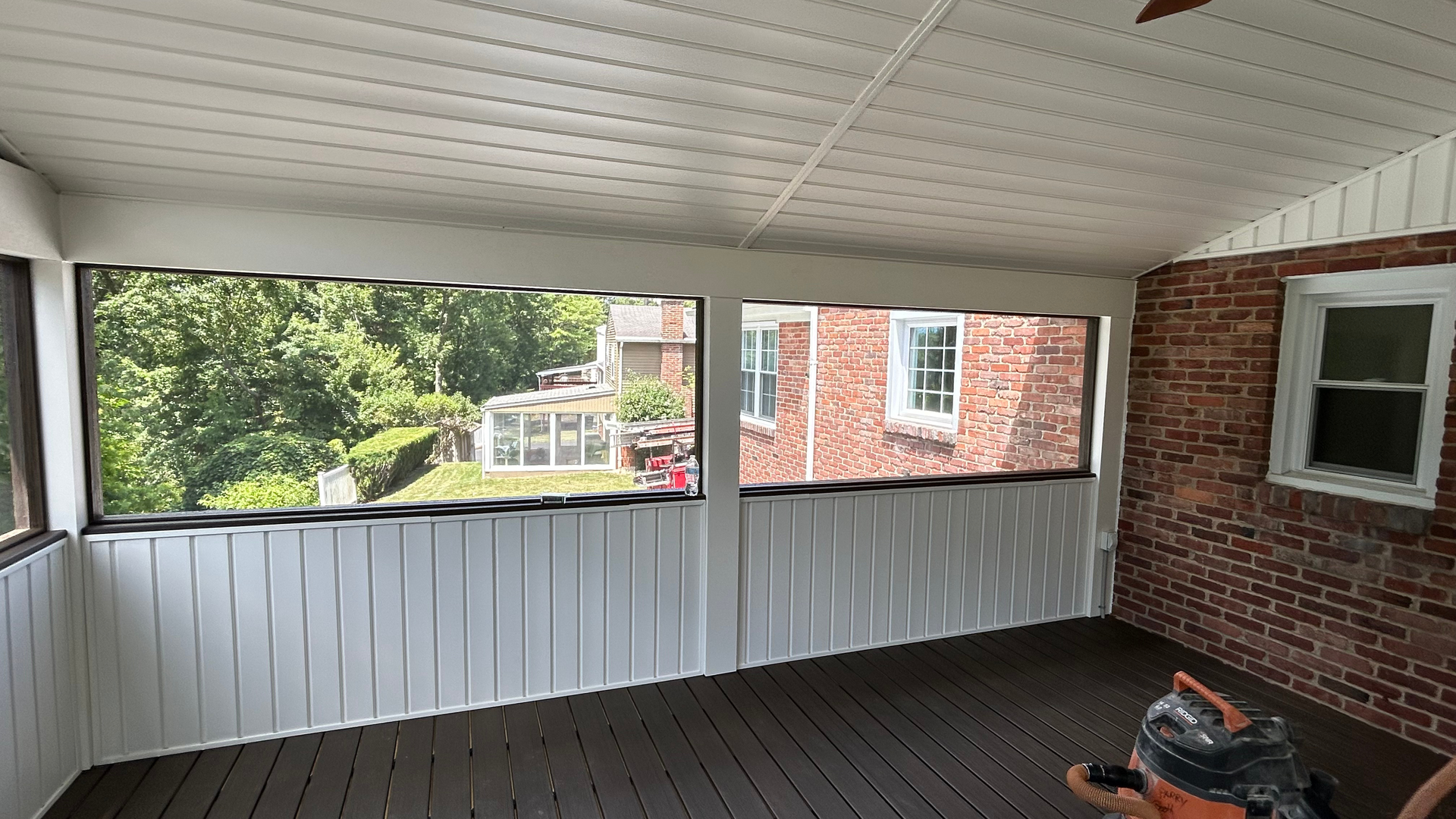 Covered porch with white ceiling, brick wall, dark floor, and a view of neighboring houses and trees.
