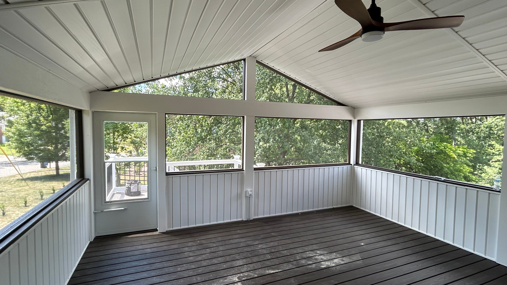 Covered porch with white railing, screened openings, dark floor, and a ceiling fan overlooking trees.