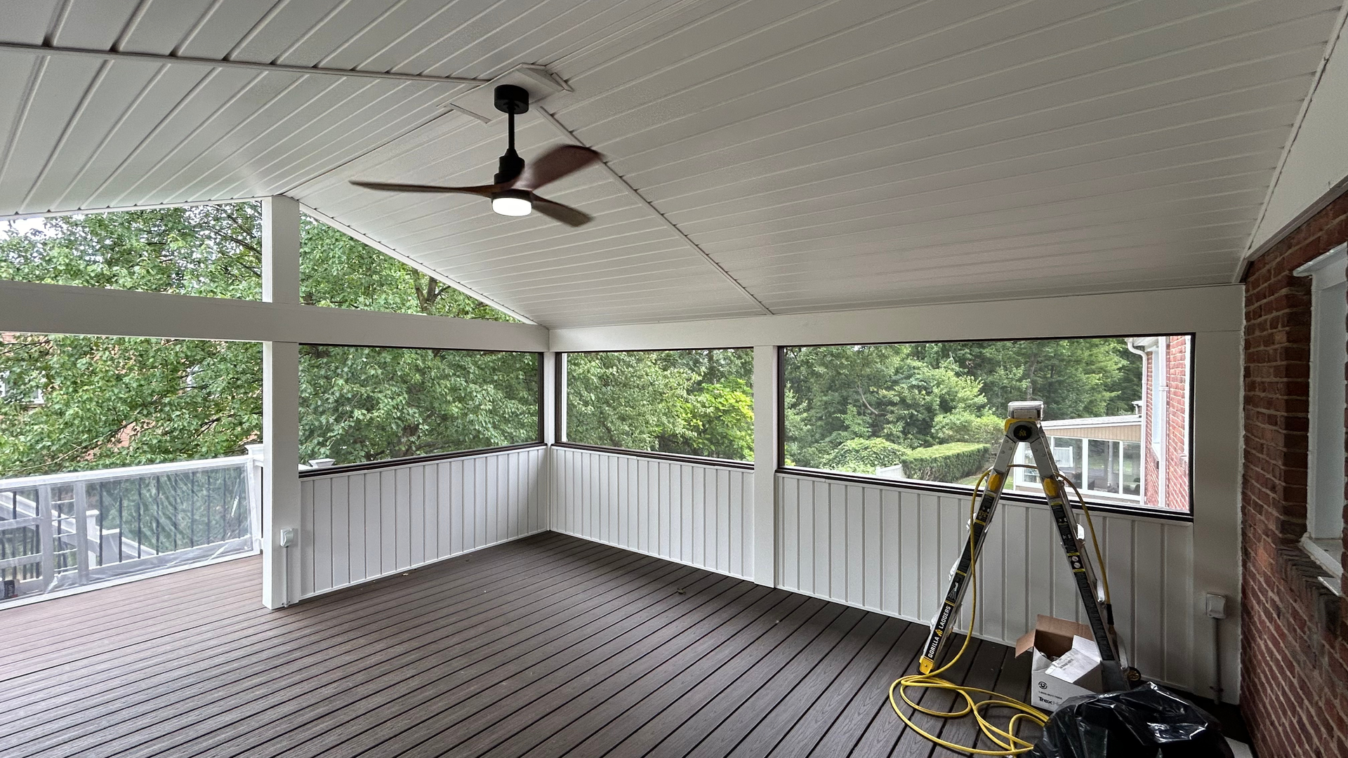 Covered porch with white ceiling fan, gray floor, railing, trees, and a ladder near the corner.