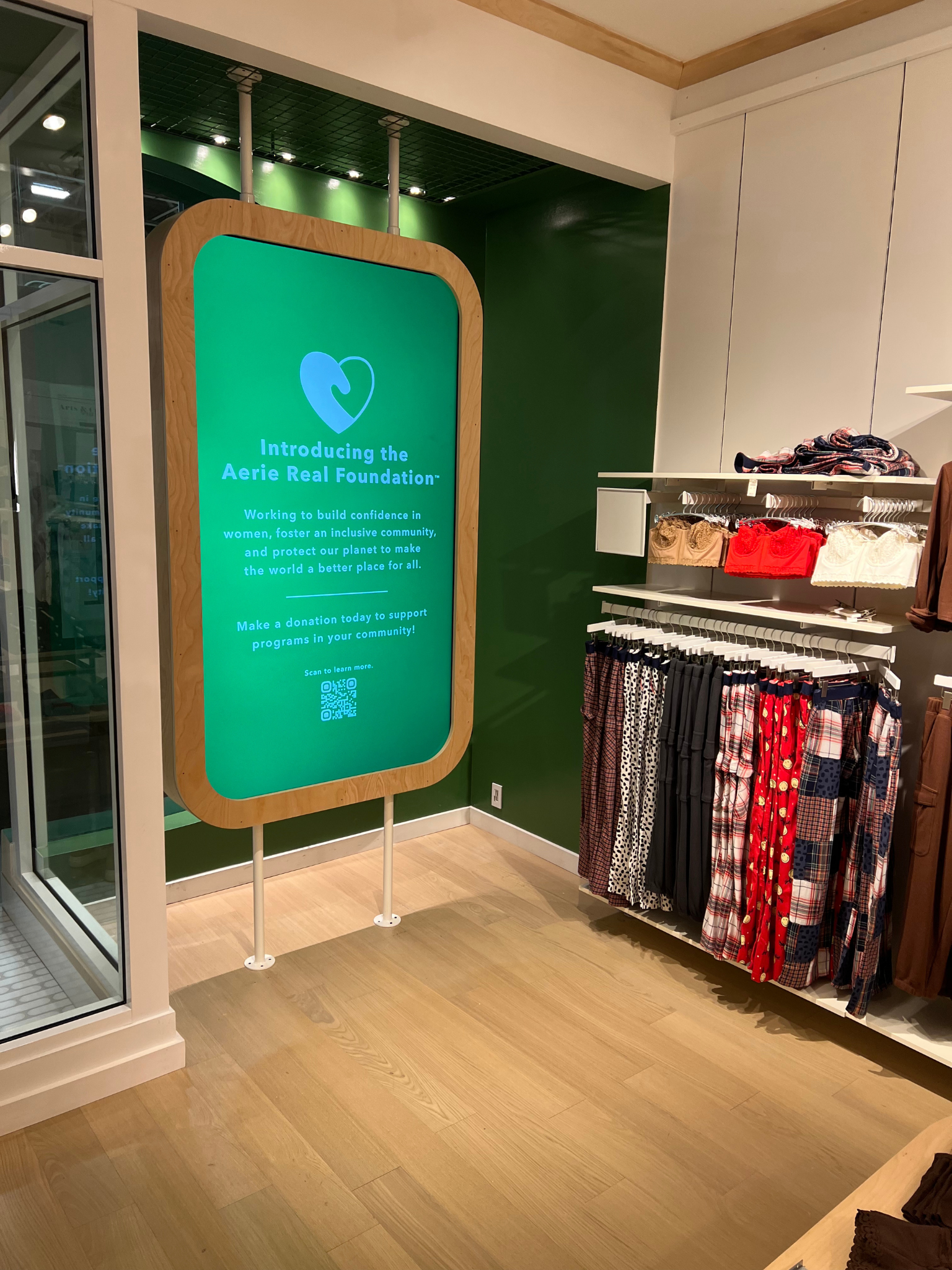 Retail store corner with a green informational sign, glass wall, and a rack of folded or hanging accessories.