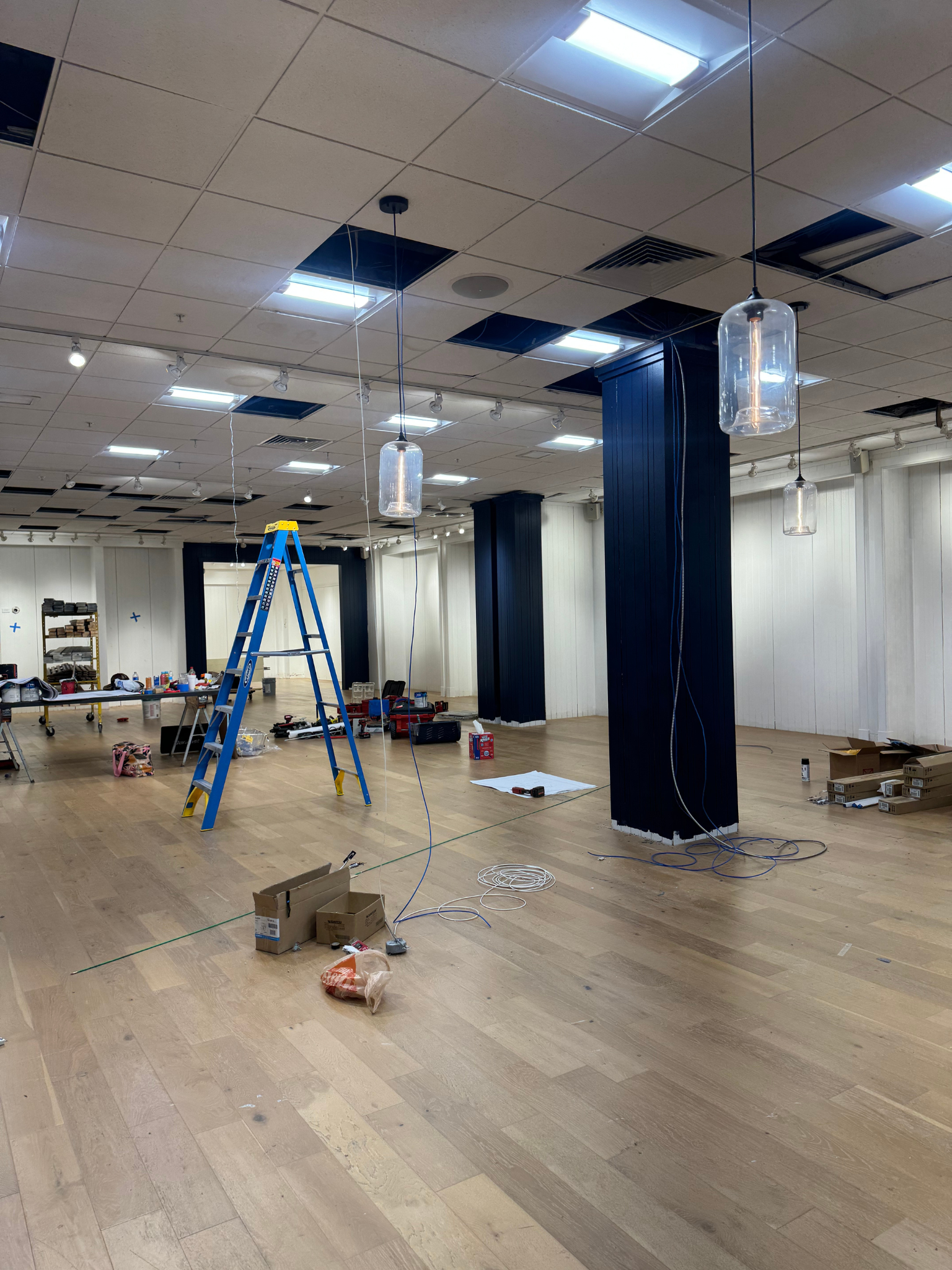Damaged empty room with broken ceiling panels, debris on the floor, and a blue ladder under fluorescent lights