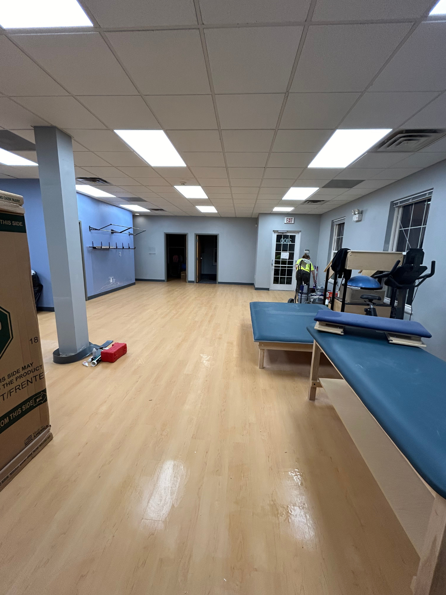 Empty physical therapy room with blue treatment tables, wood floor, mirrors, and a person near the back.