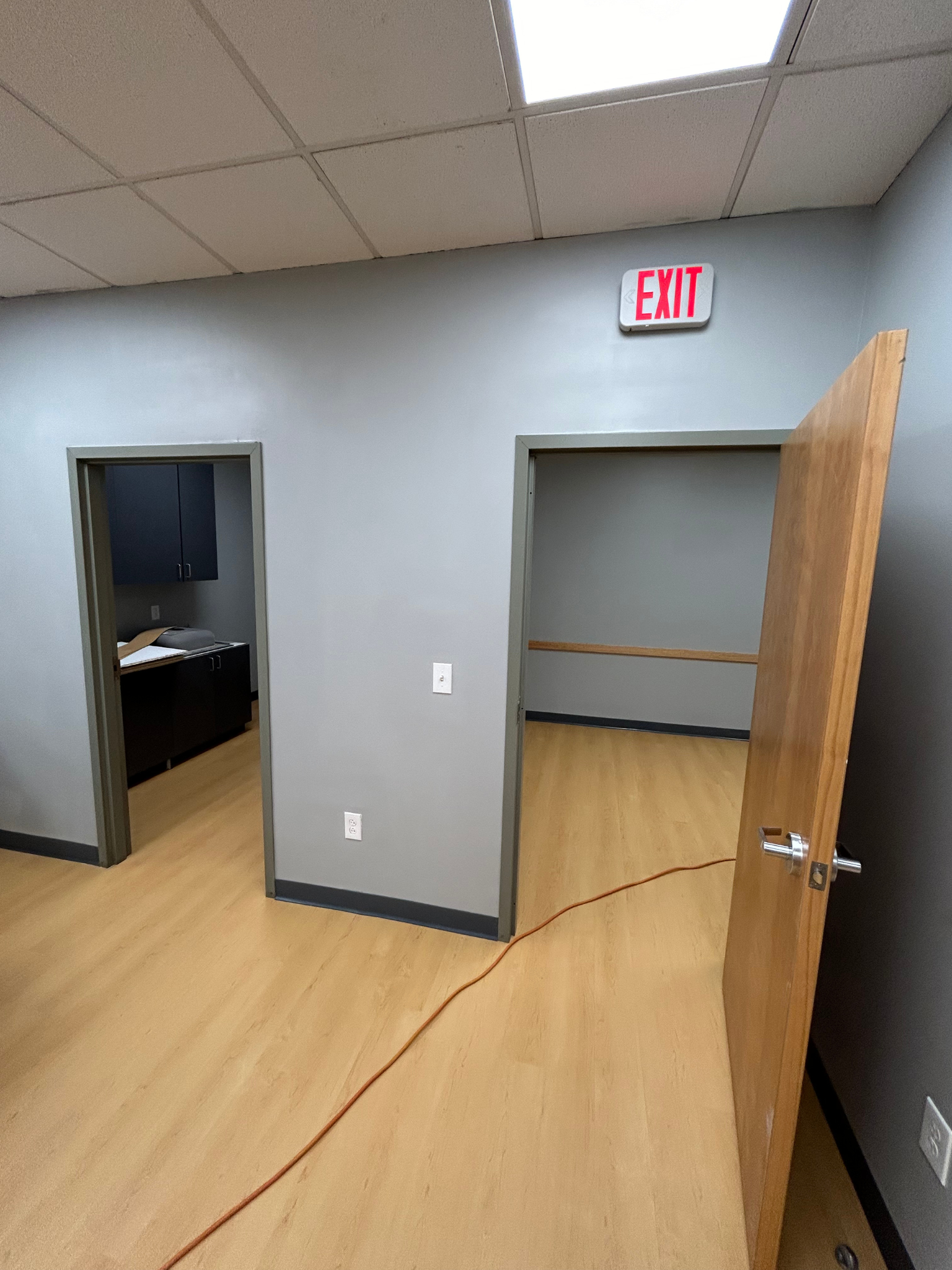 Empty office interior with open doors, wood floor, gray walls, and EXIT sign above doorway.