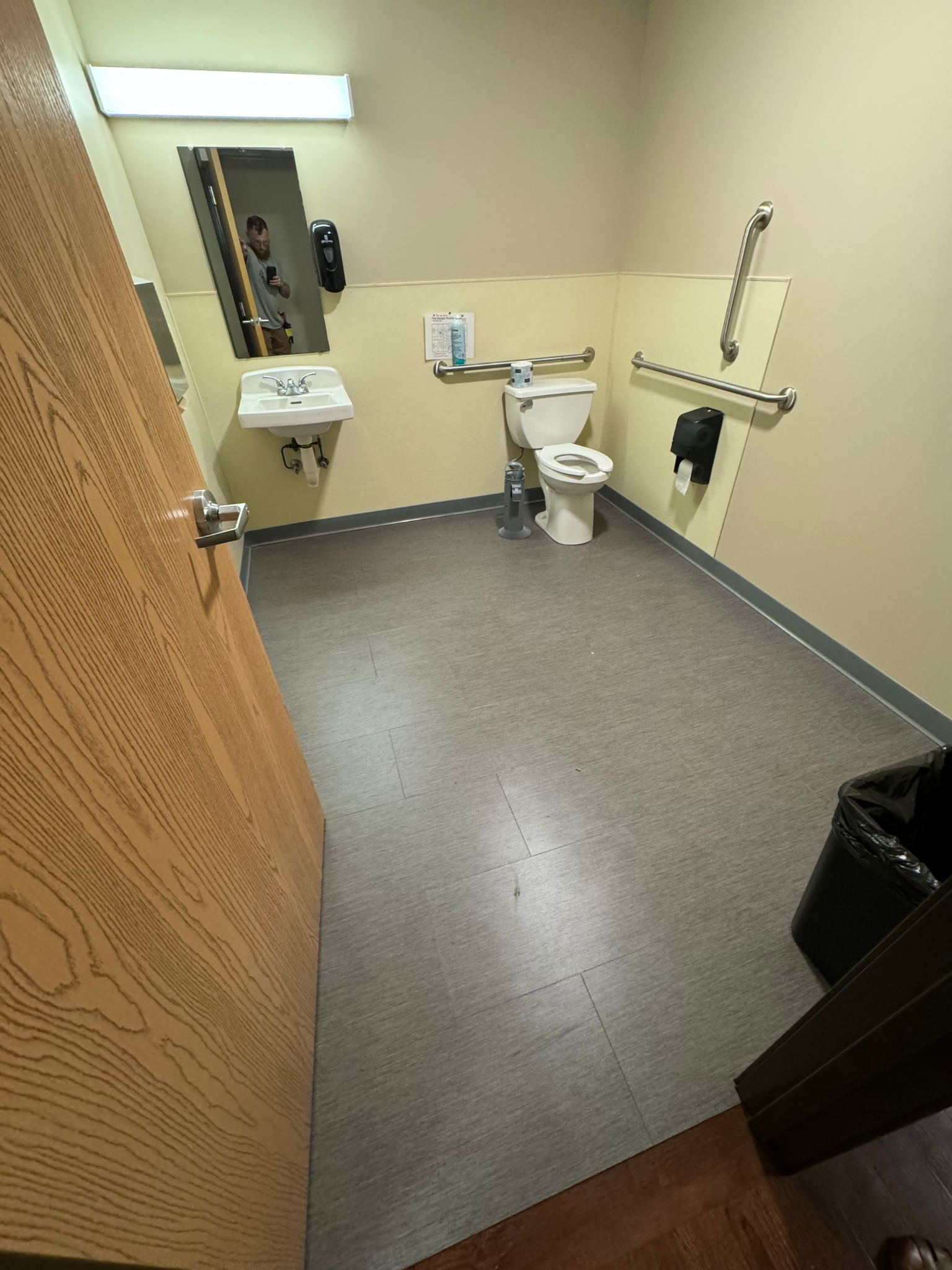 Small restroom with toilet, sink, mirror, grab bars, and gray flooring, viewed from the doorway
