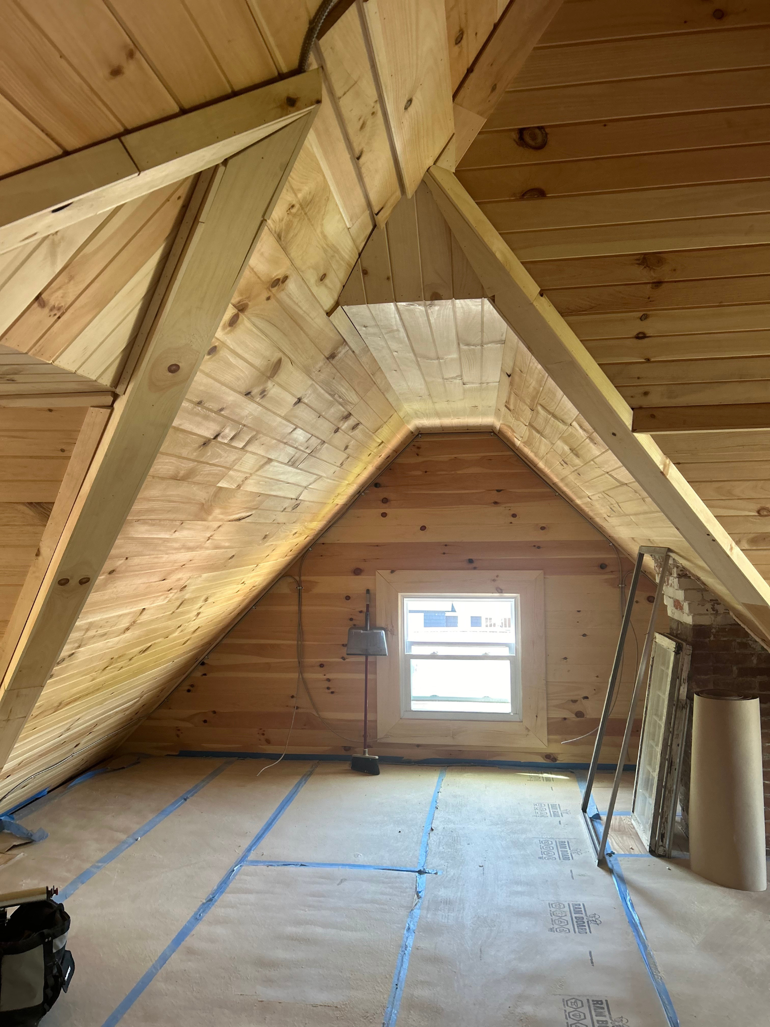 Attic room with exposed wood beams, blue tape on floor, and a small window at the far end