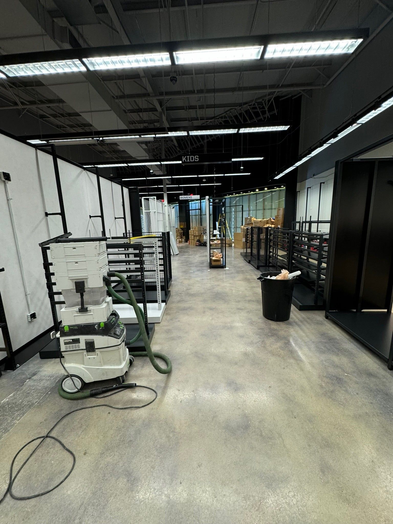 Empty industrial hallway with exposed ceiling, work lights, carts, and cleaning equipment on a concrete floor