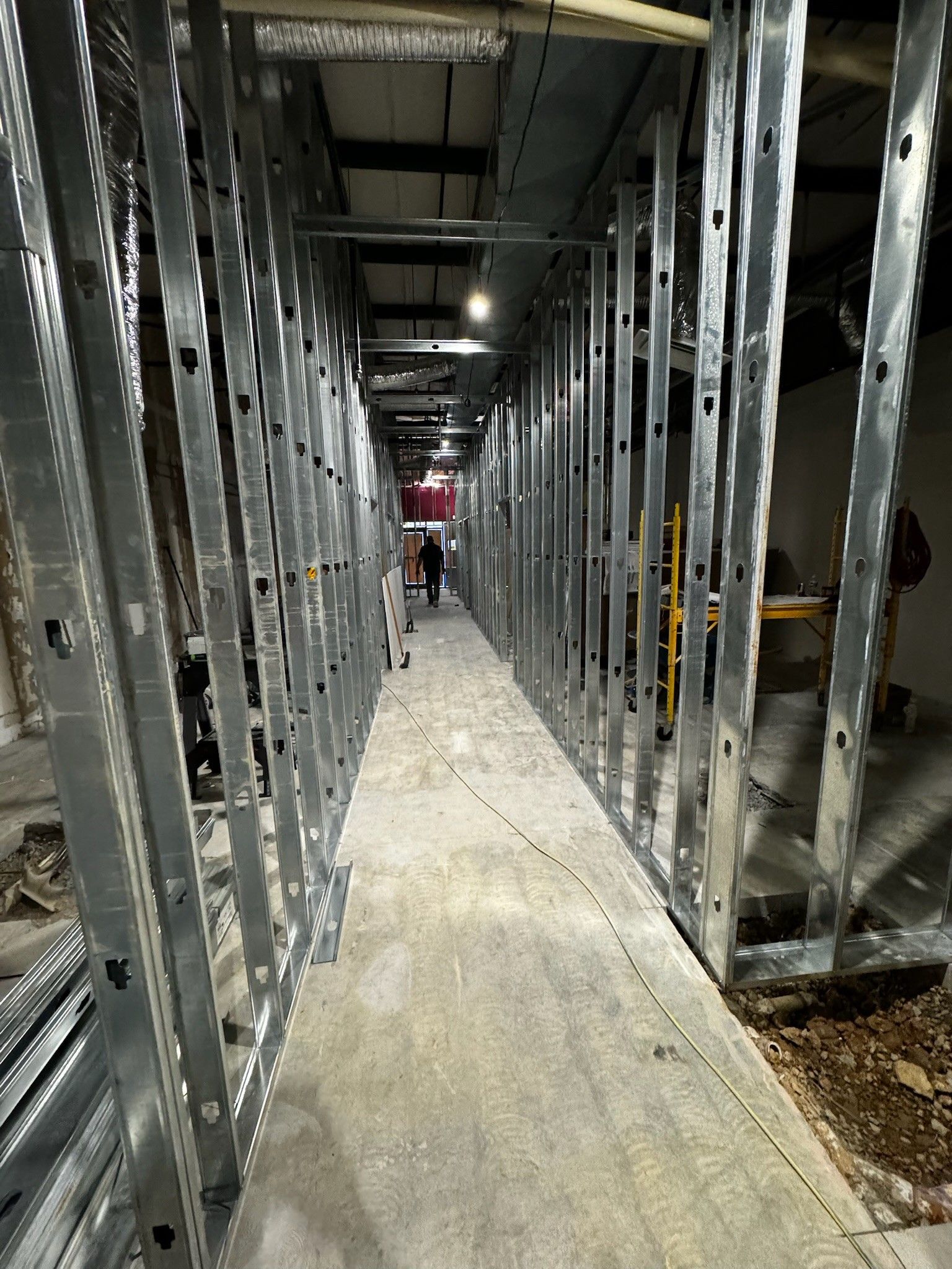 Narrow concrete corridor between exposed metal framing in an unfinished building, with a person in red at the far end